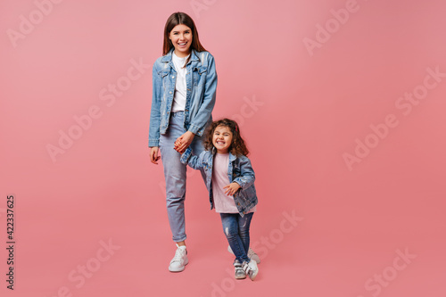 Good-humoured mom and daughter in denim jackets having fun together. Young mother and child holding hands on pink background.