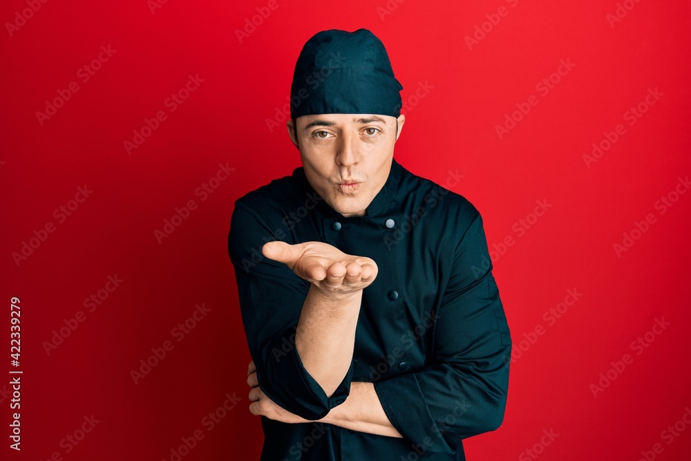 Handsome young man wearing professional cook uniform and hat looking at the camera blowing a kiss with hand on air being lovely and sexy. love expression. Handsome young man wearing professional cook uniform and hat looking at the camera blowing a kiss with hand on air being lovely and sexy. love expression.