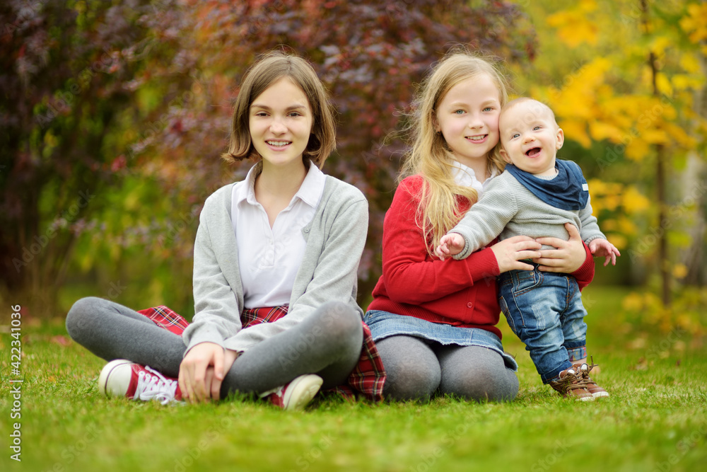 Two big sisters and their infant brother having fun outdoors. Two young girls holding their baby boy sibling on summer day. Kids with large age gap. Big age difference between siblings. Two big sisters and their infant brother having fun outdoors. Two young girls holding their baby boy sibling on summer day. Kids with large age gap. Big age difference between siblings.
