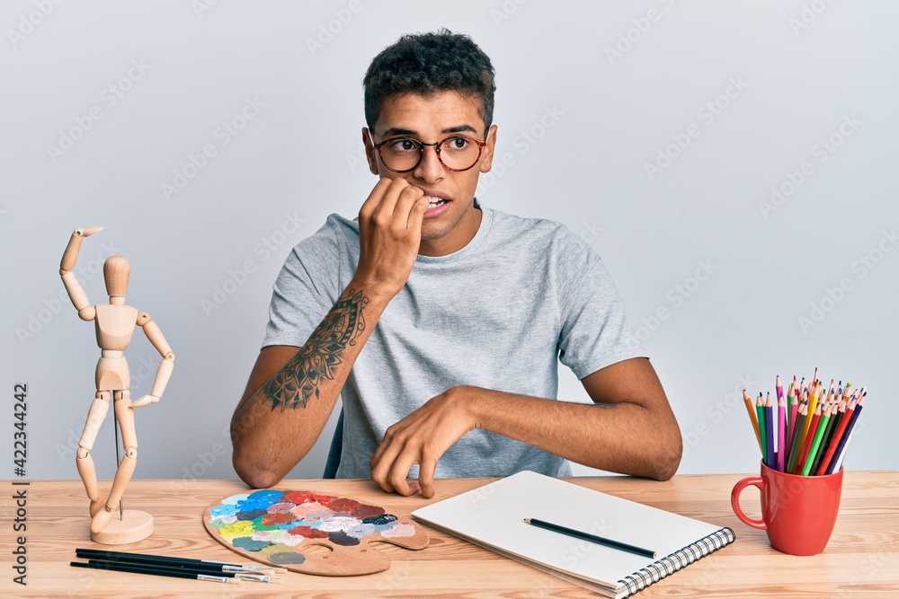 Young handsome african american man painter sitting palette and art manikin looking stressed and nervous with hands on mouth biting nails. anxiety problem. Young handsome african american man painter sitting palette and art manikin looking stressed and nervous with hands on mouth biting nails. anxiety problem.