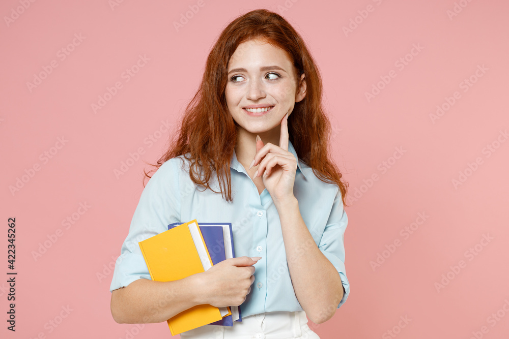 Young pensive redhead student woman in blue shirt holding books notebooks look aside prop up chin isolated on pastel pink background studio portrait. Education high school university college concept