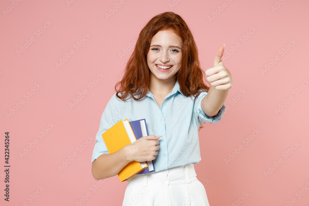 Young happy fun redhead student woman in blue shirt hold books notebook show thumb up like gesture isolated on pastel pink background studio portrait. Education high school university college concept Young happy fun redhead student woman in blue shirt hold books notebook show thumb up like gesture isolated on pastel pink background studio portrait. Education high school university college concept