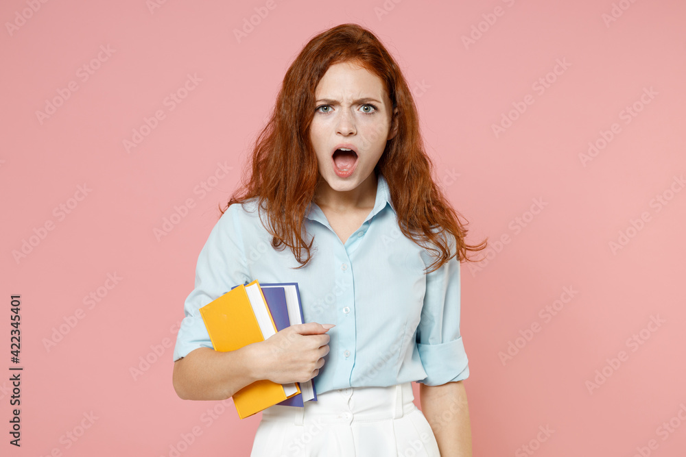 Young indignant redhead student woman in blue shirt hold book notebook look camera with open mouth isolated on pastel pink background studio portrait. Education high school university college concept Young indignant redhead student woman in blue shirt hold book notebook look camera with open mouth isolated on pastel pink background studio portrait. Education high school university college concept