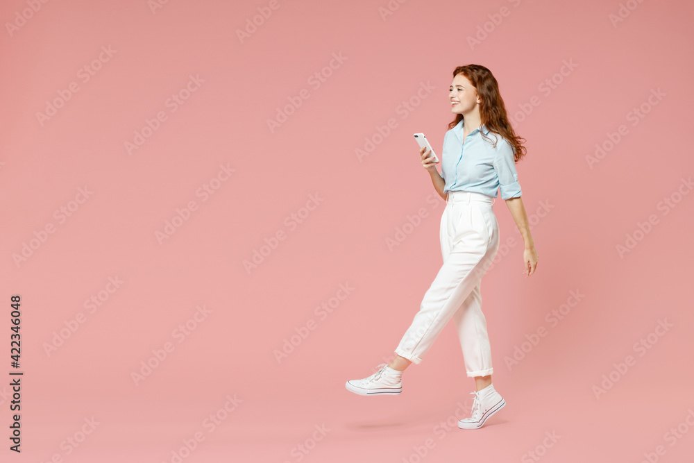 Full length side view of young student smiling redhead woman 20s in blue shirt pants using mobile cell phone chatting online in social network walk isolated on pastel pink background studio portrait.