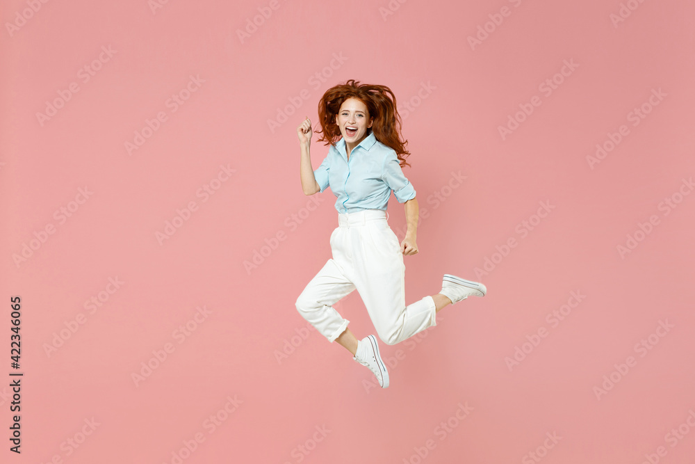 Full length young student successful overjoyed happy redhead woman 20s in blue shirt pants do winner gesture clench fist celebrate jumping high run isolated on pastel pink background studio portrait