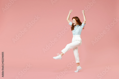 Full length body young student successful overjoyed excited happy redhead woman 20s in blue shirt pants walk do winner gesture clench fist celebrate isolated on pastel pink background studio portrait.