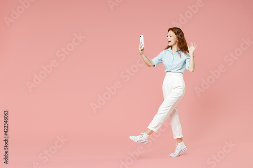 Full length side view of young friendly redhead woman in blue shirt pants doing selfie shot on mobile phone talk by video call waving hand greeting isolated on pastel pink background studio portrait.