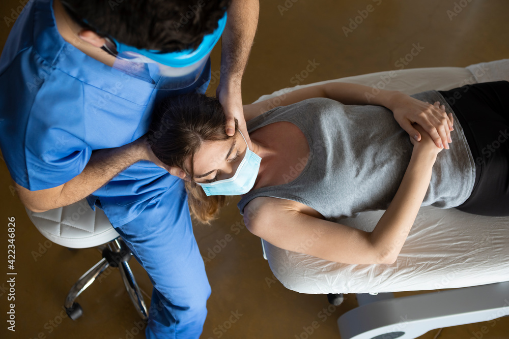 Top angle of a physiotherapist wearing a mask and uniform applying pressure to the neck of a female young patient lying on a stretcher in a clinic, health medical center during Coronavirus pandemic. Top angle of a physiotherapist wearing a mask and uniform applying pressure to the neck of a female young patient lying on a stretcher in a clinic, health medical center during Coronavirus pandemic.