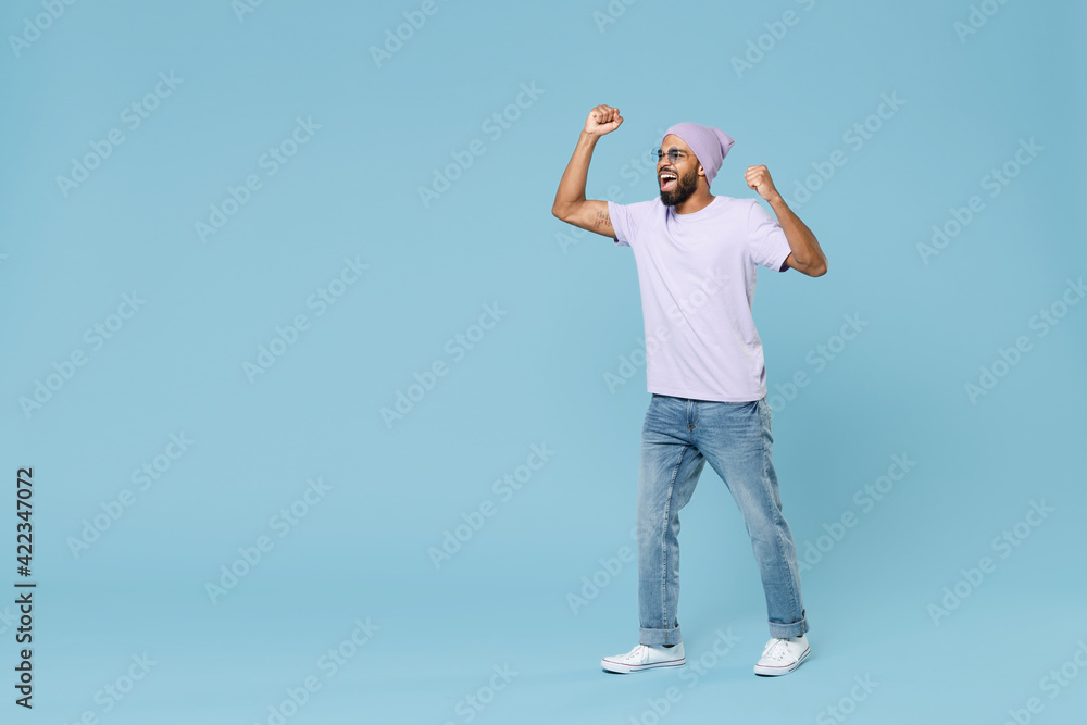 Full length young overjoyed excited fun unshaven black african man in violet t-shirt hat glasses do winner gesture clench fist celebrate scream isolated on pastel blue background studio portrait Full length young overjoyed excited fun unshaven black african man in violet t-shirt hat glasses do winner gesture clench fist celebrate scream isolated on pastel blue background studio portrait
