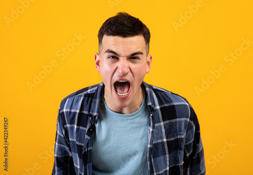 Frustrated young guy shouting in anger, expressing his fury over orange studio background. Negative emotions concept