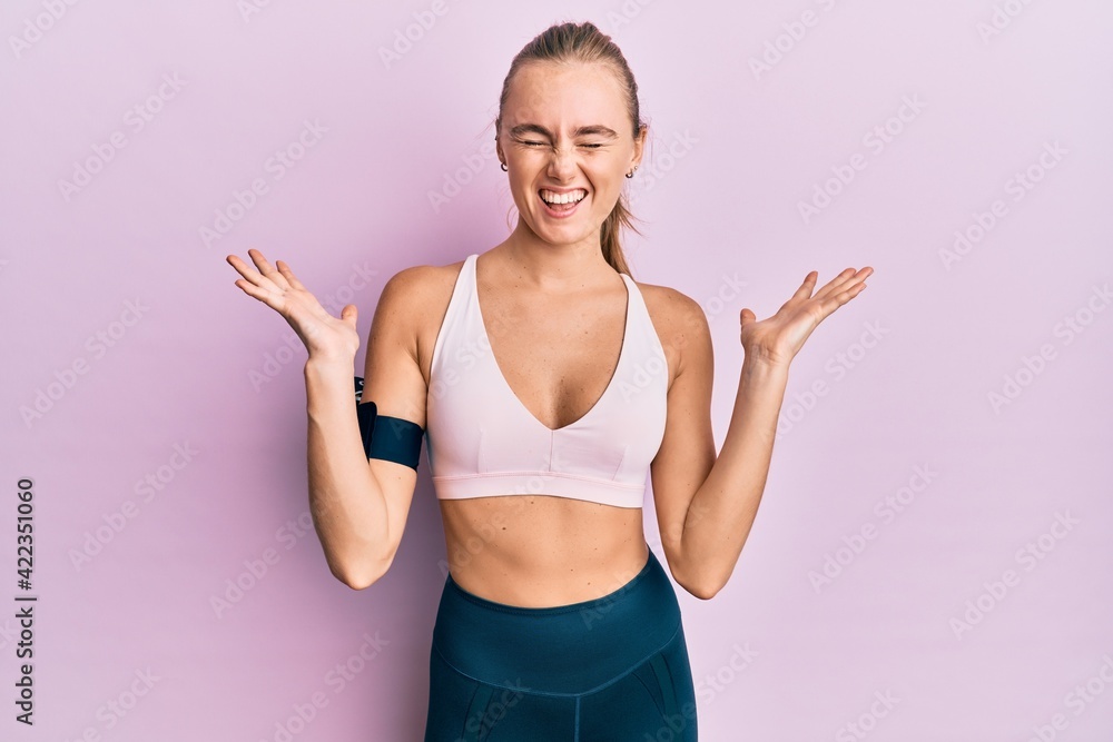 Beautiful blonde woman wearing sportswear and arm band celebrating mad and crazy for success with arms raised and closed eyes screaming excited. winner concept Beautiful blonde woman wearing sportswear and arm band celebrating mad and crazy for success with arms raised and closed eyes screaming excited. winner concept