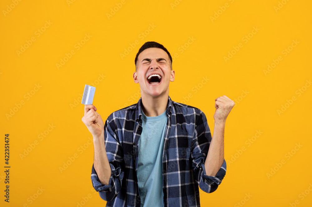 Excited young guy with credit card screaming OMG and making YES gesture over orange studio background Excited young guy with credit card screaming OMG and making YES gesture over orange studio background