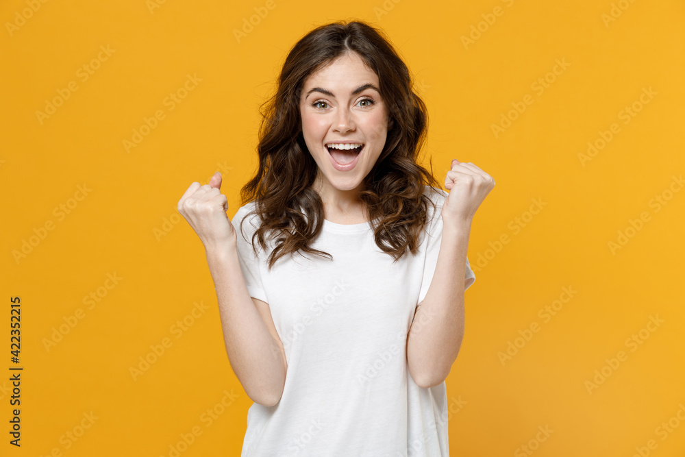 Ταπετσαρία Young smiling happy fun overjoyed caucasian student woman 20s in white basic casual blank print design t-shirt do winner gesture clench fist celebrating isolated on yellow background studio portrait