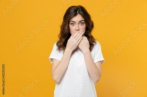 Young shocked impressed surprised amazed eueropean caucasian student woman 20s wear white basic casual t-shirt cover mouth with hand look camera isolated on yellow orange background studio portrait.