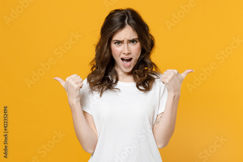 Young shocked surprised woman 20s in white casual blank print design t-shirt point thumb fingers aside on workspace area copy space mock up isolated on yellow orange color background studio portrait