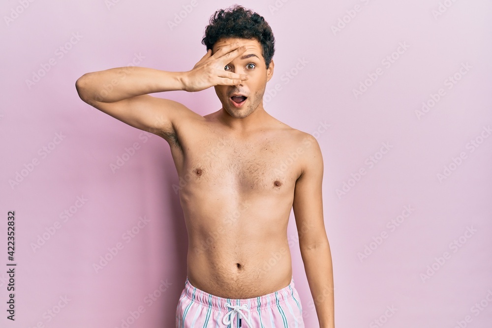Fototapeta premium Young handsome man wearing swimwear shirtless peeking in shock covering face and eyes with hand, looking through fingers with embarrassed expression.