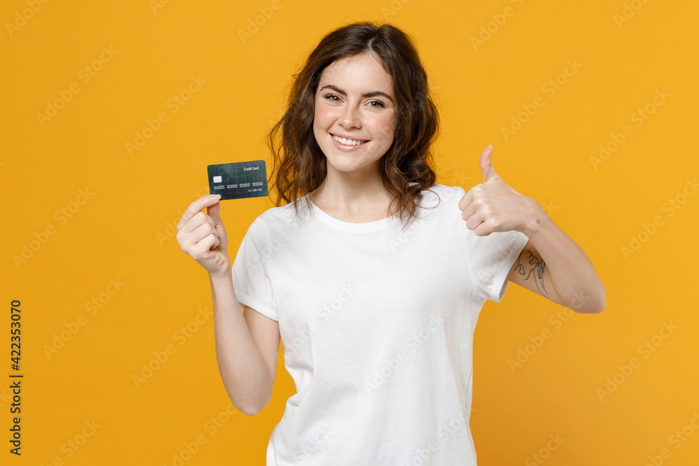 Young smiling happy rich caucasian student woman 20s wear blank print design white basic t-shirt hold credit bank card show thumb up like gesture isolated on yellow orange background studio portrait. Young smiling happy rich caucasian student woman 20s wear blank print design white basic t-shirt hold credit bank card show thumb up like gesture isolated on yellow orange background studio portrait.
