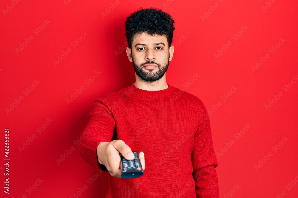 Young arab man with beard holding television remote control relaxed with serious expression on face. simple and natural looking at the camera. Young arab man with beard holding television remote control relaxed with serious expression on face. simple and natural looking at the camera.