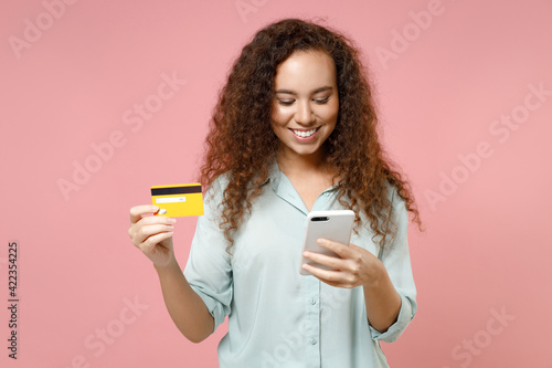 Young black african curly smiling fun woman 20s wear casual blue shirt hold in hand using mobile cell phone credit bank card shopping online isolated on pastel pink color background studio portrait