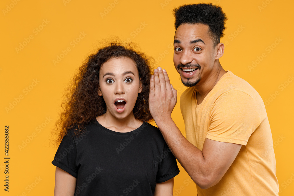 Fototapeta premium Young couple two friends family african shocked man woman in black t-shirt whisper gossip and tell secret behind hand share news with guy look camera isolated on yellow background studio portrait.