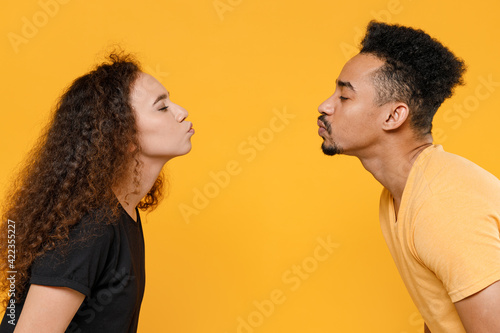 Side view of young romantic couple friends together family african woman man 20s in black t-shirt kiss each other with closed eyes while stand face to face isolated yellow background studio portrait.