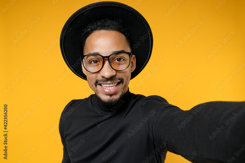 Close up of young smiling fun happy african american man 20s in stylish black hat shirt eyeglasses do selfie shot on mobile phone looking camera isolated on yellow orange background studio portrait Close up of young smiling fun happy african american man 20s in stylish black hat shirt eyeglasses do selfie shot on mobile phone looking camera isolated on yellow orange background studio portrait