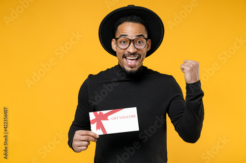 Young overjoyed american african man 20s wear stylish black shirt hat eyeglasses holding gift voucher flyer mock up do winner gesture clench fist isolated on yellow orange background studio portrait