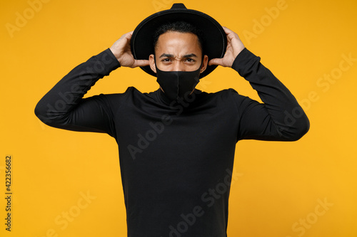 Young african confused worried man 20s in trendy stylish black hat shirt face mask to safe from coronavirus virus covid-19 pandemic quarantine hold head isolated on yellow background studio portrait. © ViDi Studio