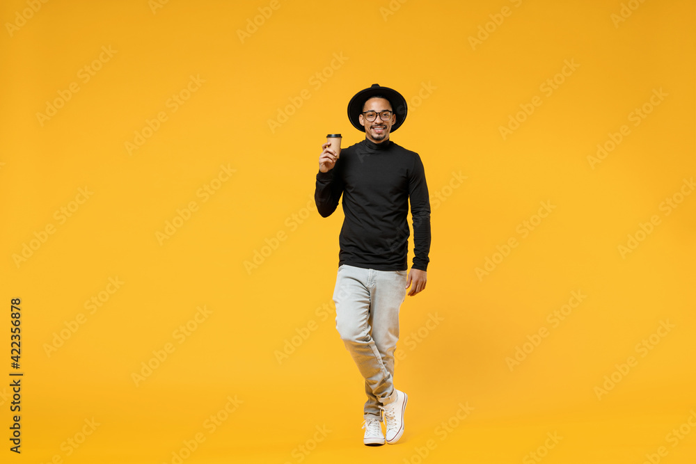 Naklejka premium Full length young happy smiling fun american african man 20s wear stylish black shirt hat eyeglasses drinking coffee hot tea in paper cup isolated on yellow orange color background studio portrait