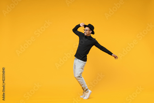 Full length young smiling fashionable fun african man 20s wearing stylish black hat shirt eyeglasses stand on toes dancing leaning back dancing isolated on yellow orange background studio portrait