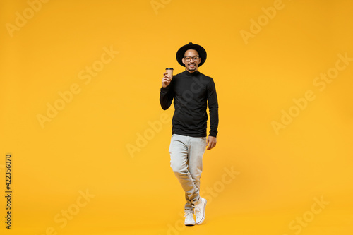 Full length young happy smiling fun american african man 20s wear stylish black shirt hat eyeglasses drinking coffee hot tea in paper cup isolated on yellow orange color background studio portrait