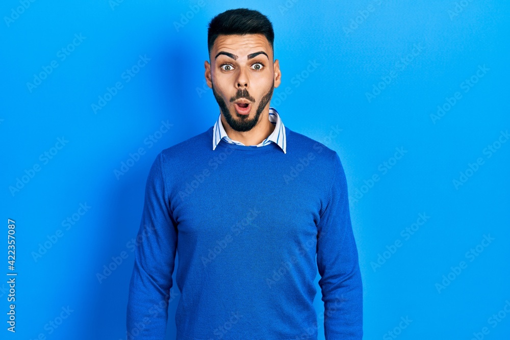 Young hispanic man with beard wearing casual blue sweater afraid and shocked with surprise and amazed expression, fear and excited face.
