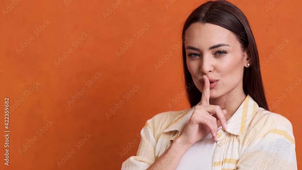 A smiling young woman is sharing secrets standing isolated over an orange background in the studio A smiling young woman is sharing secrets standing isolated over an orange background in the studio