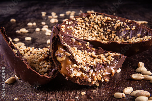 Stuffed chocolate easter egg with peanut and caramel on a wooden table with lots of peanuts around. Cracked. Close up.