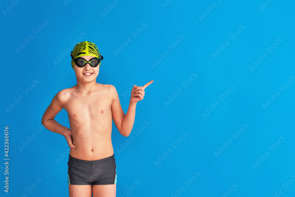 small swimmer wearing a swimming cap, glasses and a bathing suit on a blue background. child points his finger to the side, advertising. boy is studying at sports school, learning to swim. Copy space