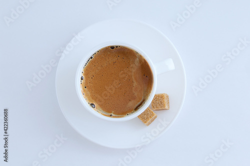 White cup of coffee with froth and cane sugar cubes on a saucer on white background.