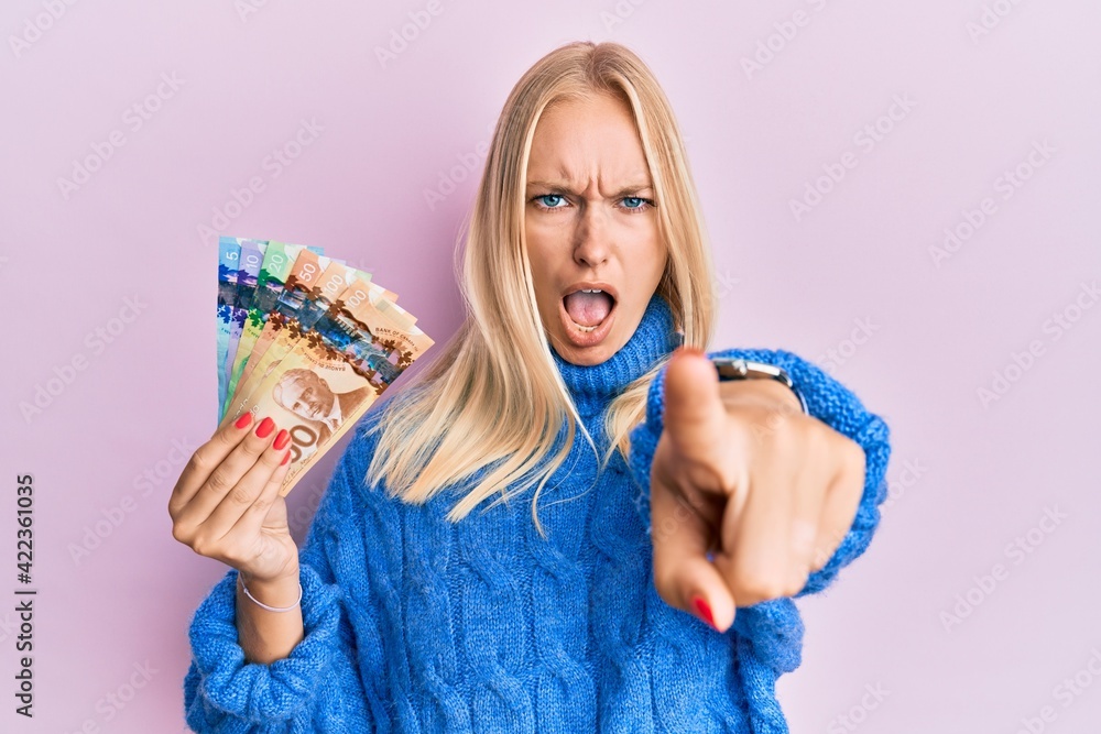 Young blonde girl holding canadian dollars pointing displeased and frustrated to the camera, angry and furious with you Young blonde girl holding canadian dollars pointing displeased and frustrated to the camera, angry and furious with you