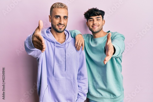 Homosexual gay couple standing together wearing casual clothes smiling friendly offering handshake as greeting and welcoming. successful business.