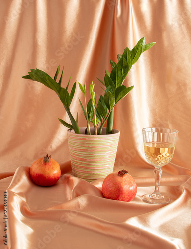 fresh green potted flowers zamia with wine glass and two fresh red pomegranate on golden curtain. creative decoration. summer background photo