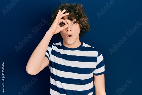 Handsome young man wearing casual striped t shirt doing ok gesture shocked with surprised face, eye looking through fingers. unbelieving expression.