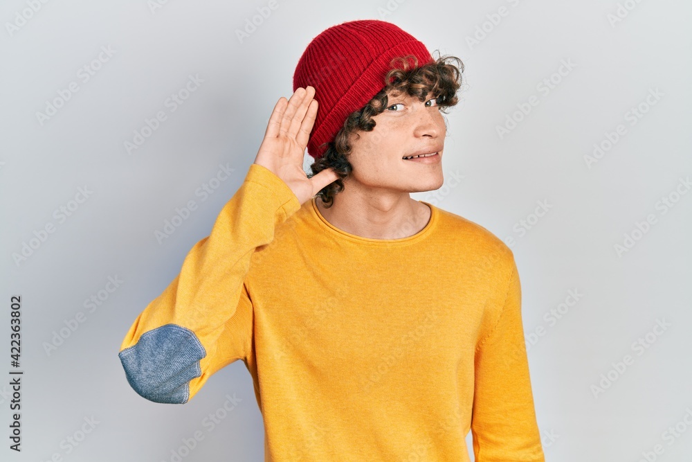Fototapeta premium Handsome young man wearing wool hat smiling with hand over ear listening an hearing to rumor or gossip. deafness concept.