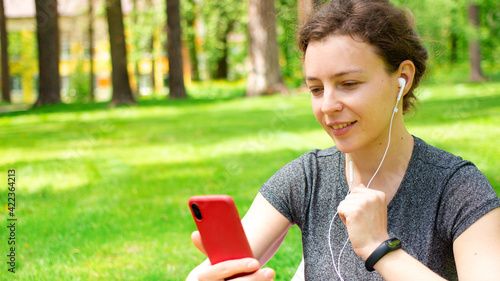 Pretty young woman looking at smartphone, sitting on green grass in city park outdoors, using headphones. Female student watching online webinar, video, listening audio course, podcast ,social media