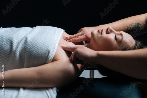 Pretty young relaxed woman lying down on massage table with closed eyes during shoulder and neck massage at spa salon. Male masseur professionally massaging shoulders on black background.