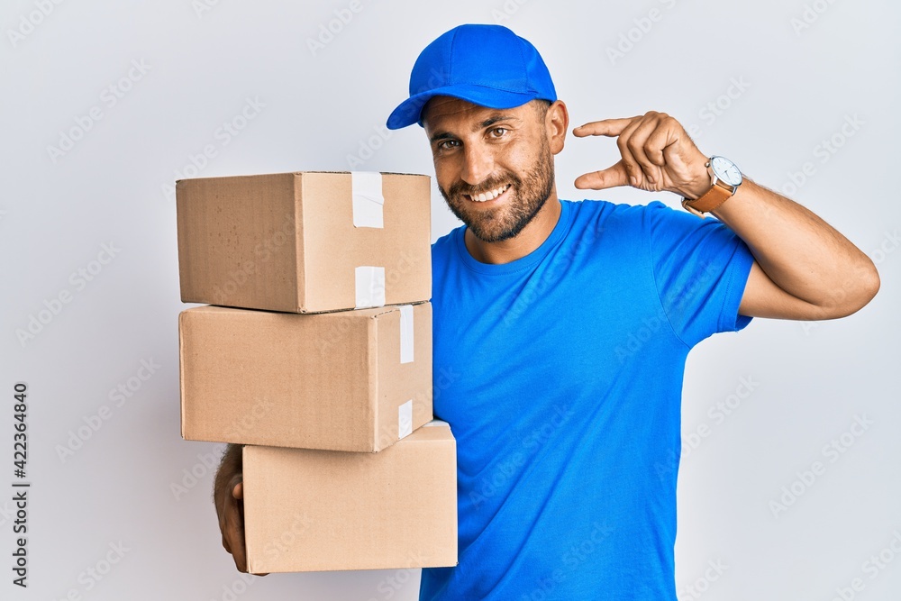 Handsome man with beard wearing courier uniform holding delivery packages smiling and confident gesturing with hand doing small size sign with fingers looking and the camera. measure concept. Handsome man with beard wearing courier uniform holding delivery packages smiling and confident gesturing with hand doing small size sign with fingers looking and the camera. measure concept.