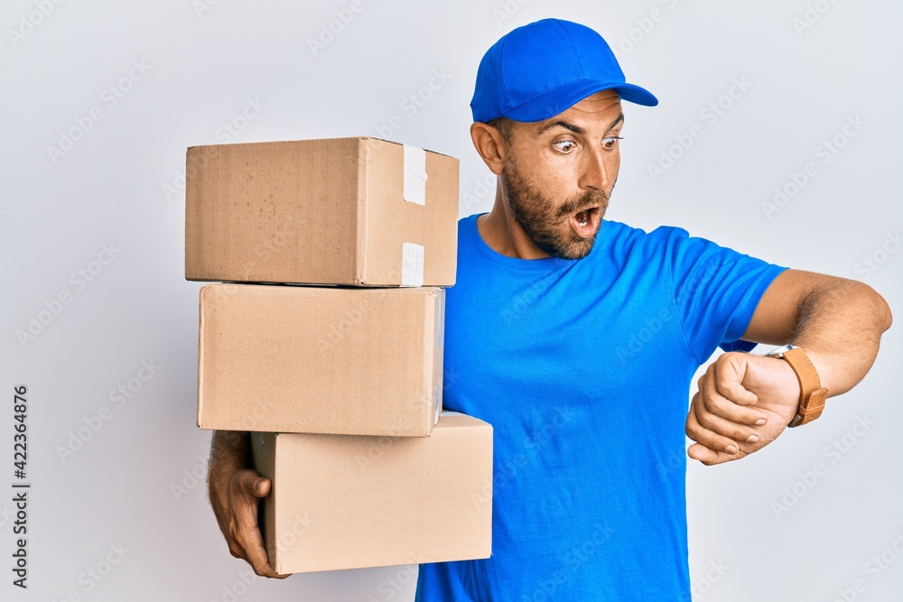 Handsome man with beard wearing courier uniform holding delivery packages looking at the watch time worried, afraid of getting late Handsome man with beard wearing courier uniform holding delivery packages looking at the watch time worried, afraid of getting late