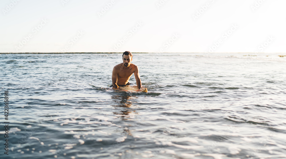 Obraz premium Alone Caucasian surfer with sexy body resting after paddle out at one of Bali top surfing beaches - Kuta, young single guy sitting at professional surfboard surrounded by ocean water on Maldives