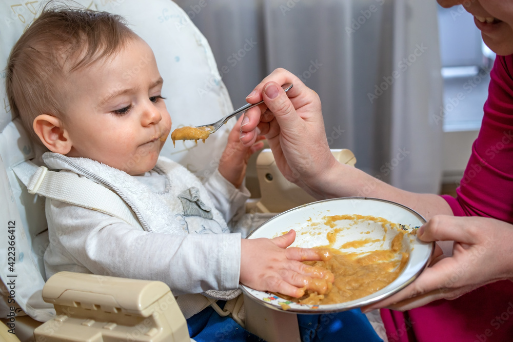 Charming messy little baby boy sitting in a feeding chair, eating and playing with his first food from the spoon with mom at home.
Charming messy little baby boy sitting in a feeding chair, eating and playing with his first food from the spoon with mom at home.