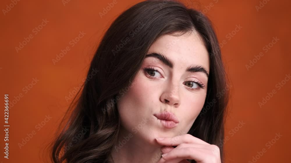 A close-up view of a beautiful young woman is thinking about something standing isolated over an orange background in the studio A close-up view of a beautiful young woman is thinking about something standing isolated over an orange background in the studio