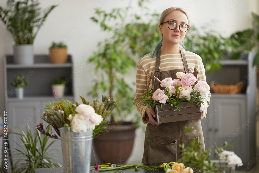 Waist up portrait of blonde young woman holding flowers and looking at camera while posing in florists workshop, copy space