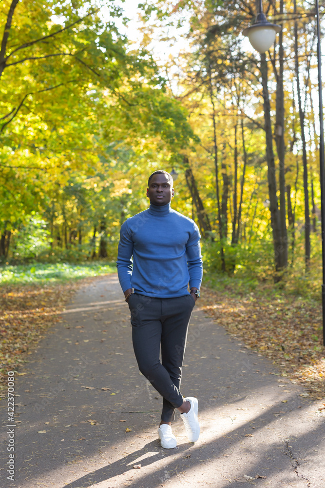 Fototapeta premium Cheerful african american young man in stylish clothes walks in autumn park on sunny warm autumn day. Concept of weekend and outdoor recreation